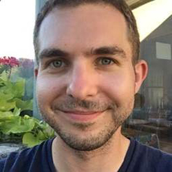 A man in a t-shirt stands outside and smiles.