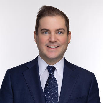 A man in a blue suit, white collared shirt, and blue tie stands and smiles