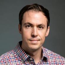 A man in a red and blue striped collared shirt stands and smiles