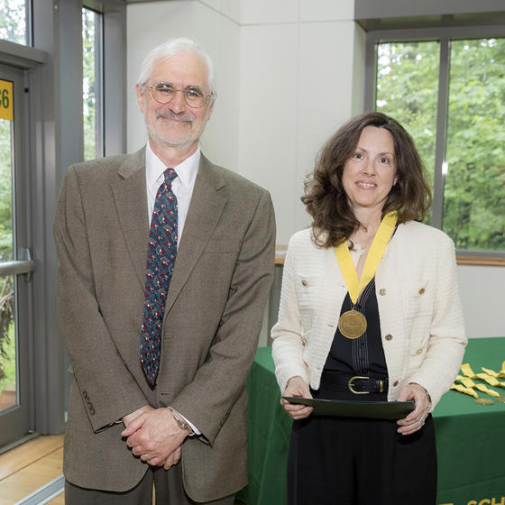 A man in a tan jacket stands next to a woman in a white jacket wearing a medal.