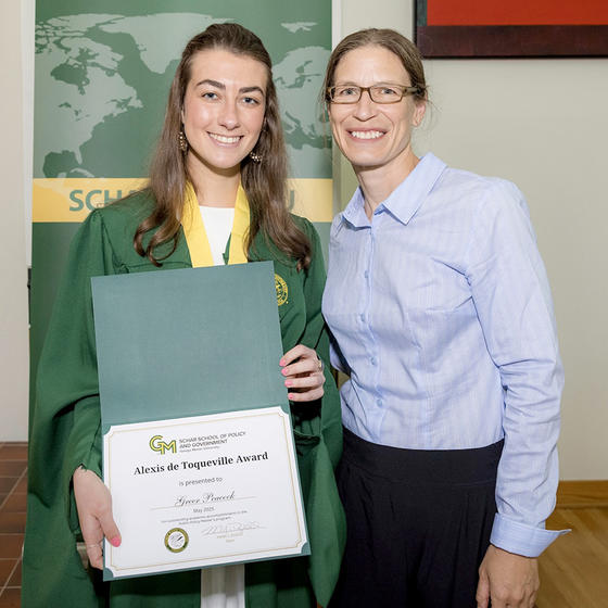 A woman in a green gown holds a certificate while a woman in a blue shirt smiles.