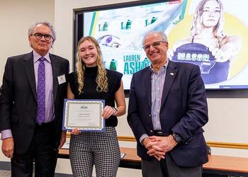 A young woman in a black top and checked slacks holds a certificate while being flanked by smiling men in suits.