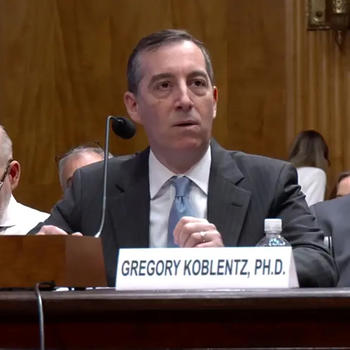 A man in a gray suit and blue tie sits at a desk with a microphone.